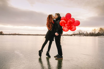 Full length of couple with balloons kissing while standing against sky