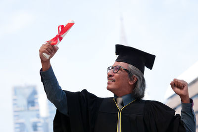 Businessman in graduation gown clenching fists against clear sky