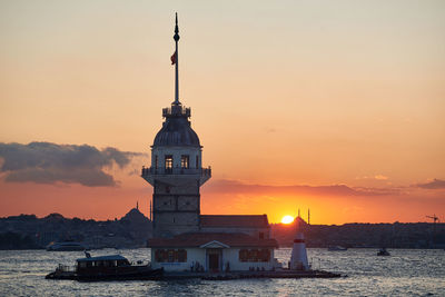 Lighthouse by sea against buildings during sunset