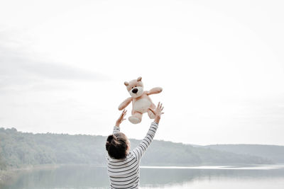 Rear view of girl playing with teddy bear by lake against clear sky