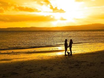Silhouette couple standing with dog on shore at beach against sky during sunset
