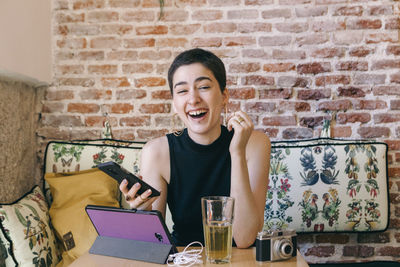 Happy young woman using phone against brick wall