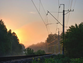 Electricity pylon by railroad tracks against sky during sunset