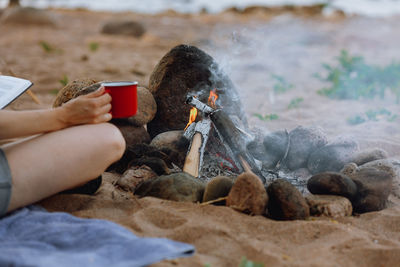 A woman's hand holds a red cup of coffee or tea against the background of a campfire