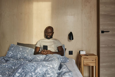 Young man using smart phone sitting on bed in bedroom at home