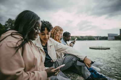 Male and female teenagers using smart phones while sitting near sea under cloudy sky