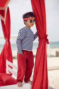 Stylish baby boy in a striped t-shirt and sunglasses stands at the red beach sunbed