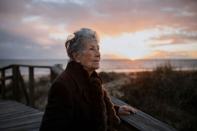 Side view of elderly female traveler in casual clothes standing on wooden pier on sandy beach and enjoying seascape at sunset