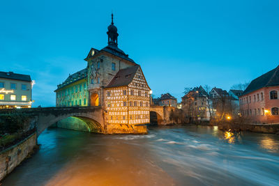 Illuminated buildings by river against blue sky