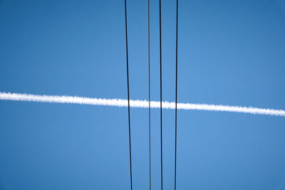 Low angle view of vapor trail and power lines against blue sky