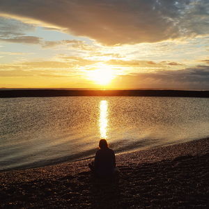 Rear view of woman sitting on sand against sea during sunset