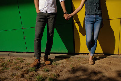 Low section of couple holding hands while standing by green and yellow wall