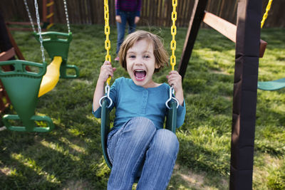 Portrait of happy girl swinging at playground with mother in background