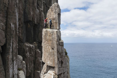 Adventurous couple summits the exposed and frail sea stack of the totem pole in a sunny day, with a sea cliff, the ocean and a sailing boat seen in the background in cape hauy, tasmania