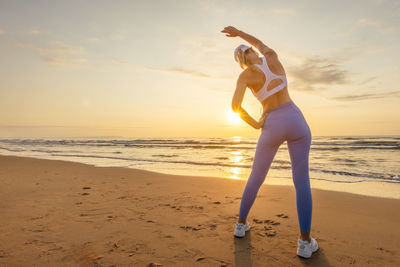 Full length of young woman exercising at beach against sky during sunset