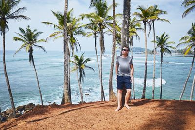 Full length of mid adult man standing by palm trees on beach against sky