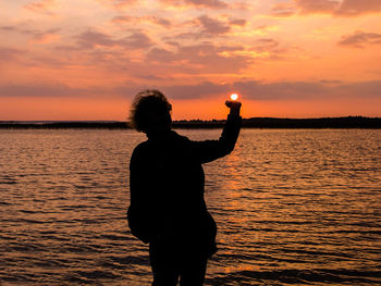 Optical illusion of silhouette person holding sun by lake during sunset