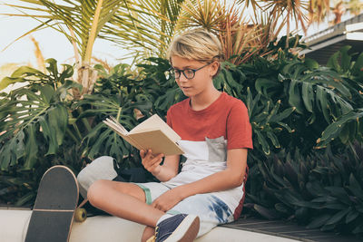 Boy reading book while sitting on seat against plants