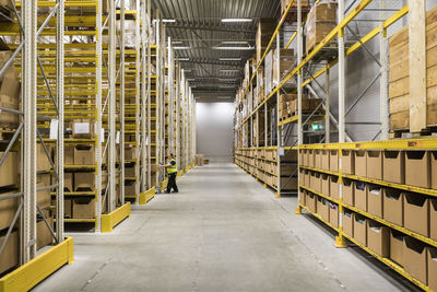 Full length of mature female warehouse worker pushing cart on aisle in industrial building