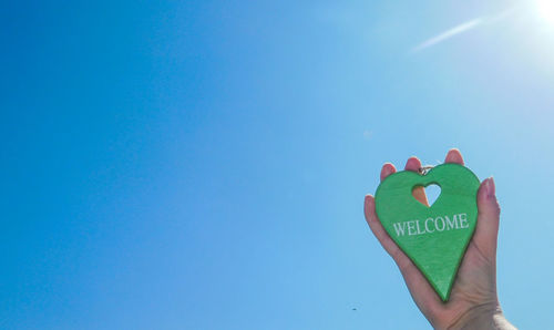 Cropped hand holding heart shape with welcome text against clear blue sky