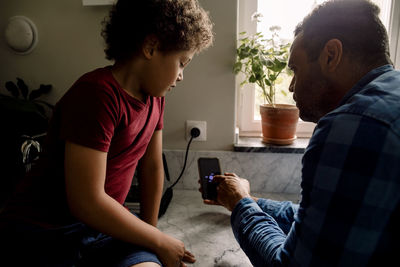 Father showing smart phone screen to son sitting on kitchen counter at home