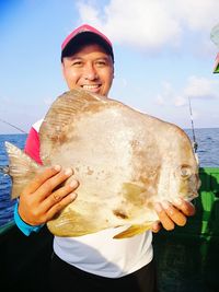 Portrait of smiling man holding dead fish while standing against sea and sky