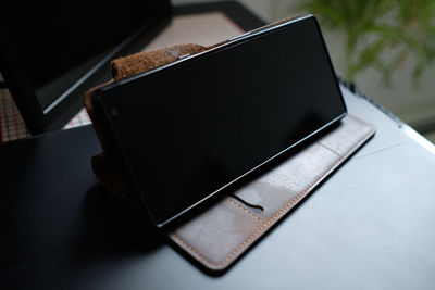 Black cellphone in a brown leather case is stood up on top of a black lap top with blank screen