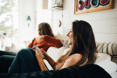 Smiling woman with smart phone looking away while sitting with friend on sofa at home