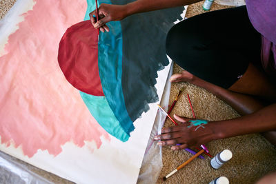 From above of crop anonymous black female with paintbrush drawing colorful picture on paper while sitting on floor at home