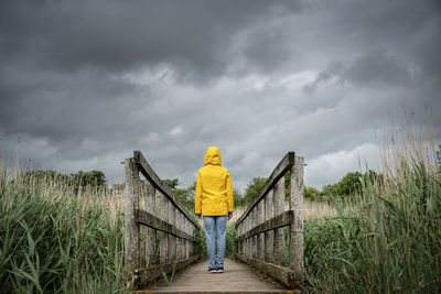 Rear view of a person standing in the middle of a wooden bridge with a dark moody sky