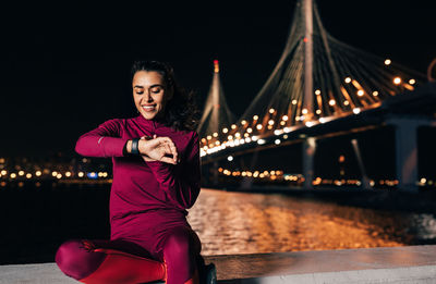 Smiling woman checking time while sitting against illuminated bridge