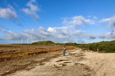 Woman walking in a dunescape under a blue sky towards a dune with wind turbines along it