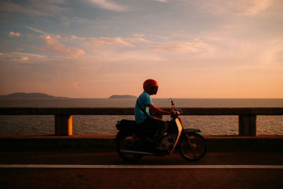 Man riding motor scooter by sea on bridge during sunset