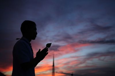 Side view of silhouette man with hands clasped praying against dramatic sky during sunset