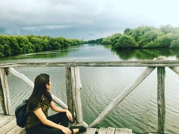 Woman sitting on railing by lake against sky