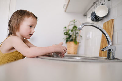 European child of four years old in yellow jumpsuit washes his hands in real bright kitchen