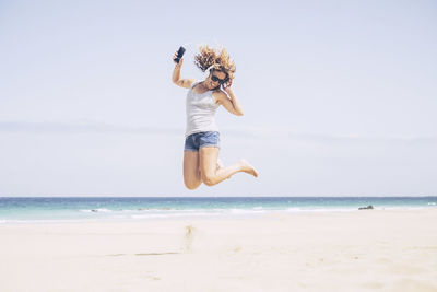 Happy young woman listening music while jumping at beach against sky during sunny day