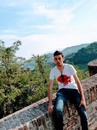 Young man sitting on tree against sky