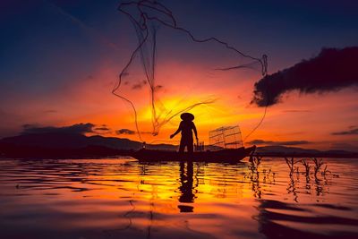 Silhouette fisherman standing in lake against sky during sunset