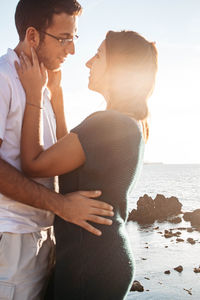 Photo of a couple looking at each other romantically standing in front of a sunset over the sea