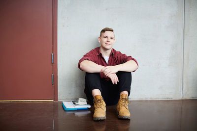 Full length portrait of male student sitting on floor against wall in university