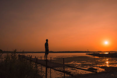 Rear view of silhouette man looking at sea against dramatic sky during sunset