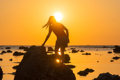 Silhouette woman standing on rock at beach against sky during sunset