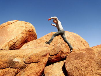 Low angle view of man jumping on rocks against clear blue sky