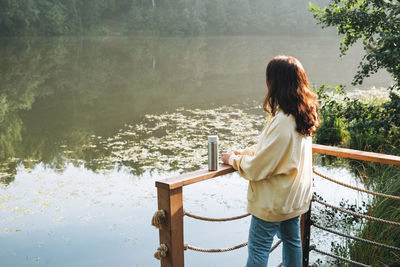 Brunette woman in yellow hoodie with thermo mug relaxing near lake in early morning
