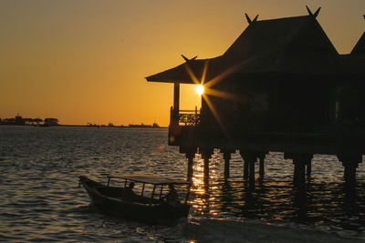 Boat sailing by stilt house in sea against clear sky during sunset