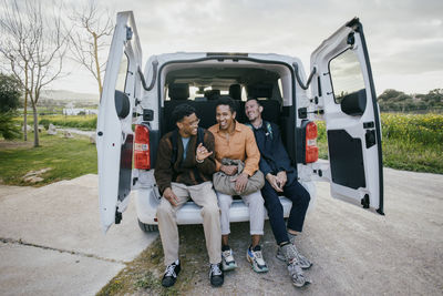 Full length of cheerful male friends sitting in van trunk on roadside