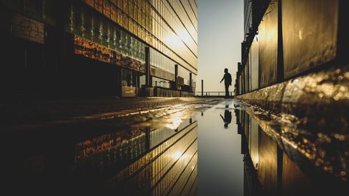 Silhouette man amidst buildings reflecting on puddle in city during sunset