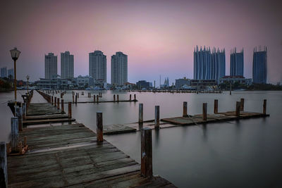Pier on river by buildings against sky during sunset
