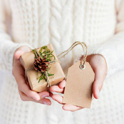 Woman in white knitted sweater holds christmas present packed in craft paper with pine cone and tag. 